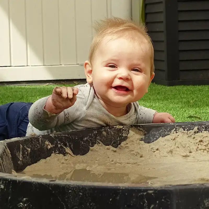 spelende-baby-zandbak-lente-de-ark-oss » Kinderopvang & BSO in Oss Baby speelt ontdekkend met zand in de tuin van Kindercentrum de Ark tijdens een zonnige lentedag in Oss.