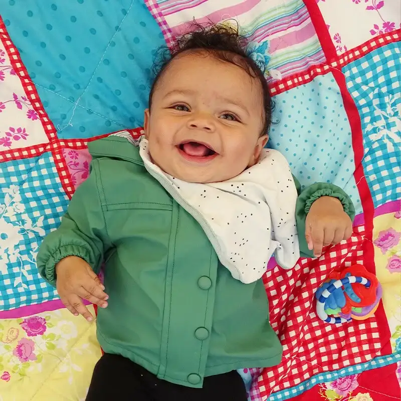 lachende-baby-buiten-speelkleed-ark-oss » Kinderopvang & BSO in Oss Een lachende baby ligt op een speelkleed buiten in het gras bij de babyopvang van Kindercentrum de Ark.