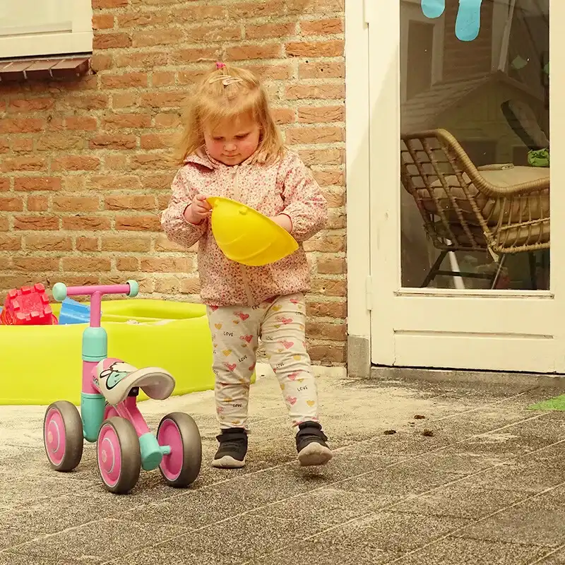 kind-met-bouwhelm-en-fietsje-buitenspelen-ark-oss » Kinderopvang & BSO in Oss Een jong kind onderzoekt een gele bouwhelm bij een loopfietsje in de tuin van Kindercentrum de Ark tijdens een lentedag in Oss.