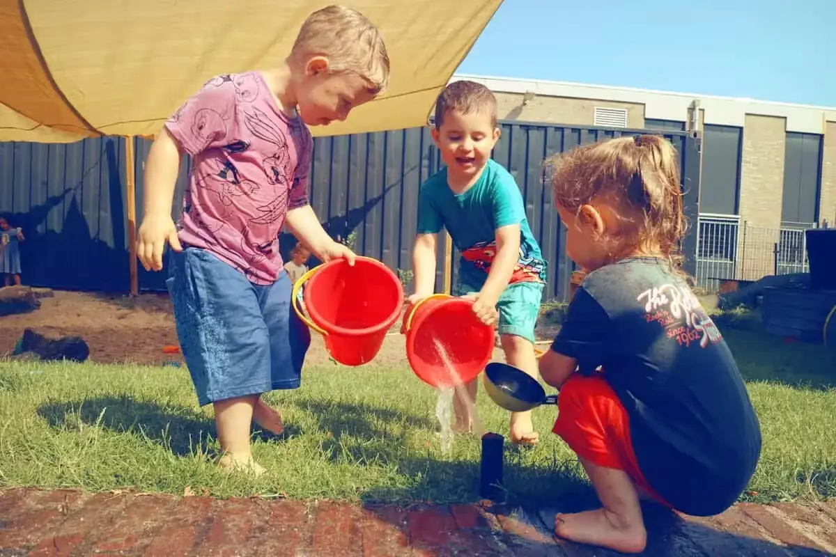 Drie jonge kinderen spelen buiten met rode emmers en water in de tuin van Kindercentrum de Ark.