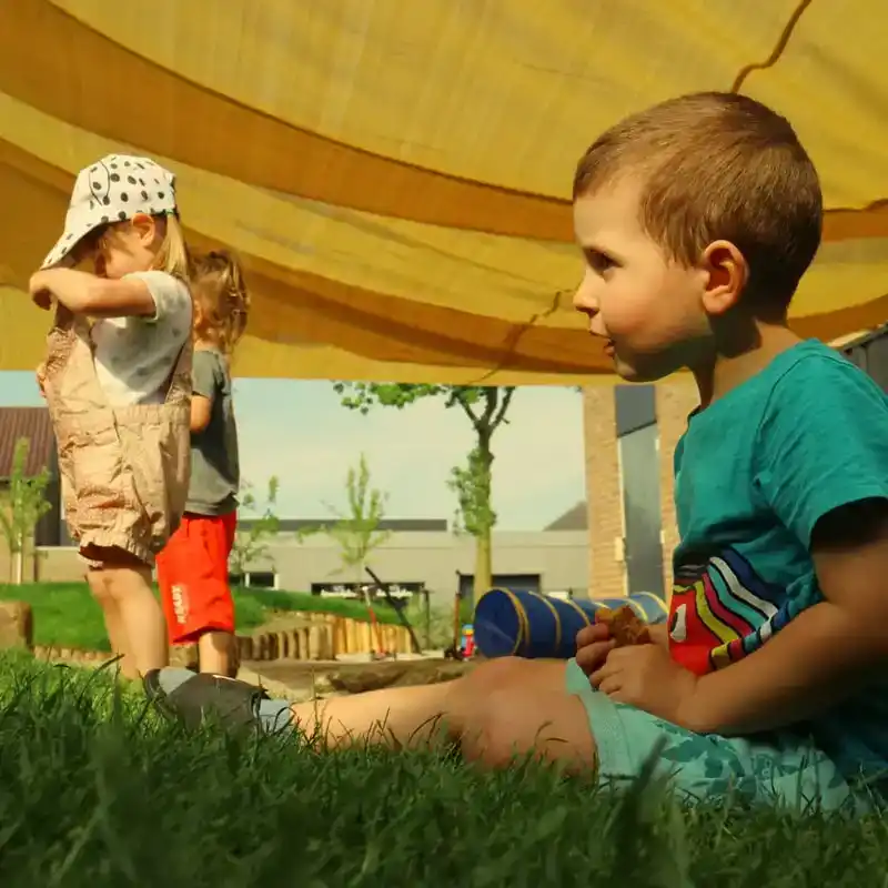 kind-speelt-in-gras-onder-schaduwdoek-ark-oss » Kinderopvang in Oss Een kind speelt rustig in het gras onder een schaduwdoek in de tuin van Kindercentrum de Ark in Oss.
