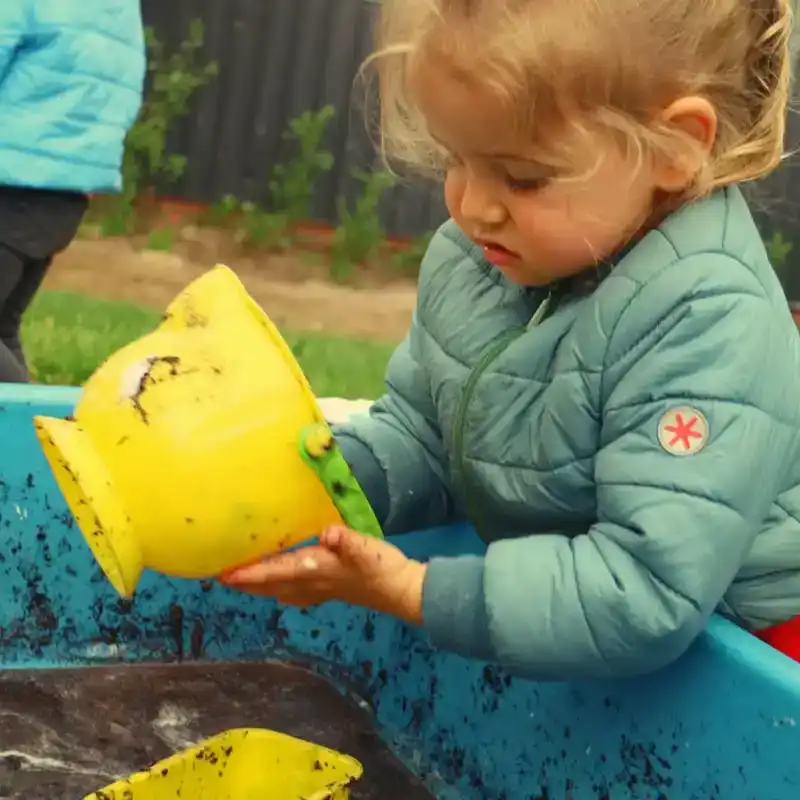 peuter-speelt-met-modderbak-ark-oss » Kinderopvang in Oss Een peuter speelt met een gele emmer bij de modderbak in de ontdektuin van Kindercentrum de Ark in Oss.